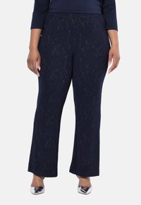 Person som bär mörkblå strukturera vida byxor och silverfärgade spetsiga skor, med händerna avslappnat vid sidorna.