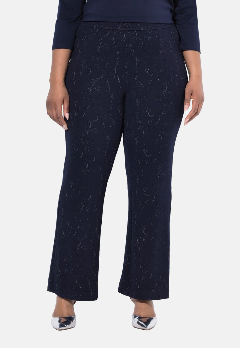 Person som bär mörkblå strukturera vida byxor och silverfärgade spetsiga skor, med händerna avslappnat vid sidorna.