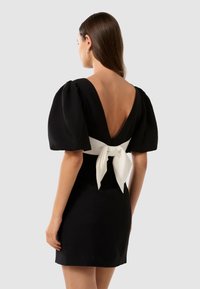 Abito nero con maniche a sbuffo e profondo scollo a V sul retro, caratterizzato da un dettaglio di fiocco bianco in vita. Tessuto morbido, forma aderente.
