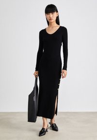 Femme portant une longue robe noire ajustée avec des boutons dorés sur la fente latérale, tenant un sac fourre-tout noir et portant des talons pointus noirs.