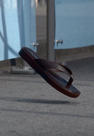 Sandale tong en cuir brun foncé suspendue en l'air au-dessus d'un pavement gris avec des cloisons bleues et des poteaux métalliques en arrière-plan.