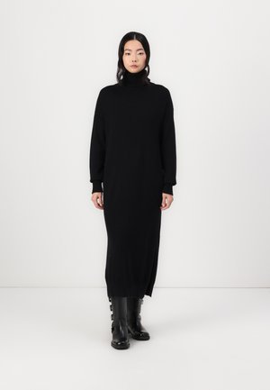 Melna adīta turtlenecka kleita ar garām piedurknēm un sānu šķēlumiem, apvienota ar melniem zābakiem līdz ceļgaliem, kas aprīkoti ar jostu akcentiem.