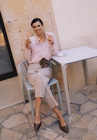 Femme souriante assise à l'extérieur sur une chaise, tenant une tasse blanche et un biscuit, à côté d'une table blanche avec une petite assiette et une cuillère.