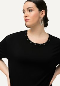 Camiseta negra con cuello redondo que presenta detalles de remaches en un lazo. La tela suave y el corte relajado mejoran la comodidad y el estilo.