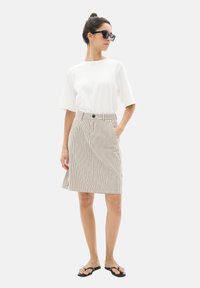 T-shirt blanc à manches courtes associé à une jupe A-line beige rayée, avec des boutons, des poches latérales et des tongs noires.