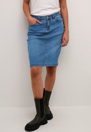Person trägt einen knielangen blauen Jeansrock, weißes Oberteil und dunkelgrüne Stiefel bis zur Mitte der Wade, steht vor einem schlichten Hintergrund.