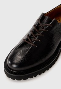 Black leather shoe with a textured finish, round toe, and brown laces. Features a thick rubber sole with a pronounced tread pattern.