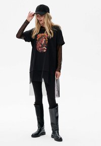 Zwarte oversized t-shirt met een kleurrafische graphic, gelaagd met een transparant zwart topje, gecombineerd met zwarte leggings en hoge zwarte laarzen.