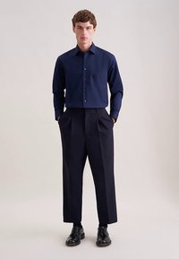 Marineblauw shirt met lange mouwen, knoopsluiting en borstzak, gecombineerd met hooggetailleerde, geplooide donkere broek en zwarte schoenen.