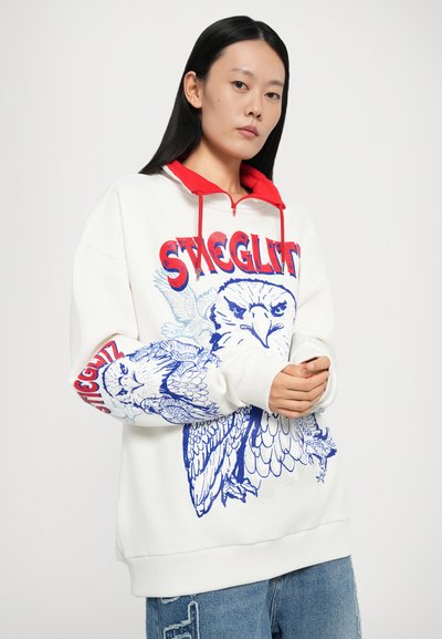 Felpa bianca con grafiche di gufo blu e accenti rossi, colletto a mezza zip e vestibilità oversize. Il testo presenta "STAGGLITZ" in rosso in grassetto.