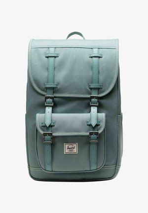 Mochila rectangular de color azul verdoso con solapa y bolsillo frontal, asegurada por dos correas de cuero con hebillas de metal. Pequeño asa en la parte superior.