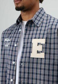 Geruit overhemd met knopen in blauw en bruin met witte stiksels en een grote stoffen "E" patch op de borst. Gemaakt van katoen, regular fit.