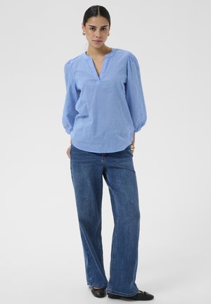 Femme aux cheveux foncés portant un chemisier bleu texturé, un jean bleu à jambes larges et des ballerines noires, debout, les mains dans le dos.