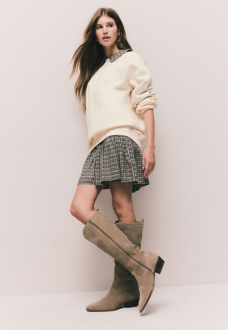 Jeune femme portant un pull beige, une jupe écossaise et des bottes en daim taupe montant jusqu'aux genoux, debout devant un mur uni de couleur claire.