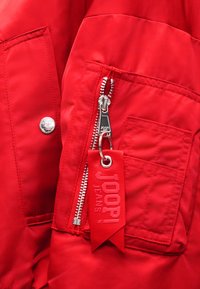 Κοντινό πλάνο στην τσέπη κόκκινης ζακέτας με ασημένιο φερμουάρ και κόκκινη ετικέτα υφάσματος με την επιγραφή "JOOP! JEANS".