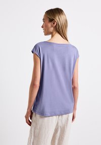 Levandulový blouse s krátkým rukávem, kulatým výstřihem a volným střihem. Hladká látka a mírně zakřivený lem. Kombinované s světle barevnými lněnými kalhotami.