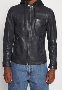 Svart läderjacka med dragkedja fram, fast huva och två sidofickor. Har en slät, jämn yta och kontrasterande detaljer.