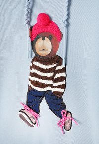 Világoskék kapucnis pulóver, amelyen egy hímzett medve látható csíkos barna-fehér pulóverben, farmernadrágban és rózsaszín fűzős cipőben, pomponos sapkával.