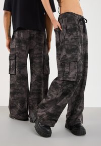 Two people wearing black and gray camouflage cargo pants, one facing forward with hand in pocket, the other facing backward, both in black shoes.