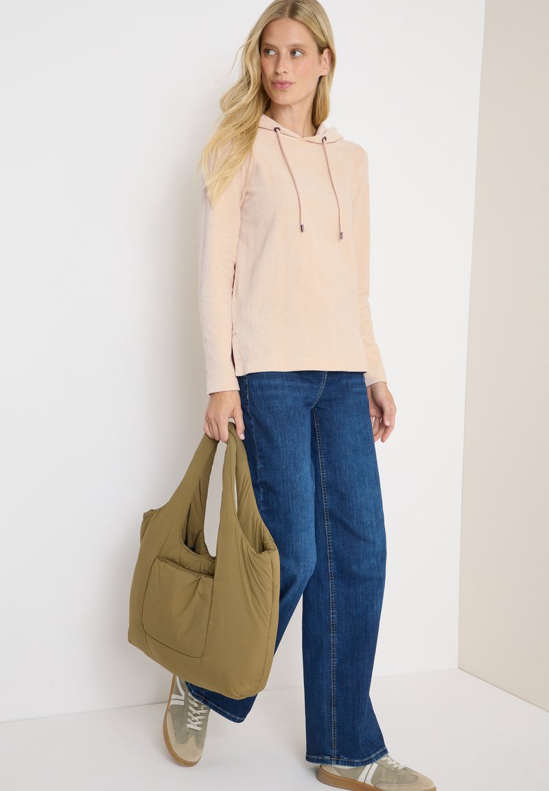 Femme portant un hoodie rose et un jean bleu, tenant un grand sac fourre-tout olive, se tenant devant un mur blanc et beige.