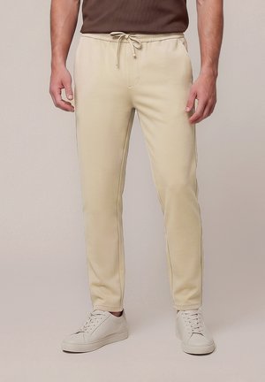 Mand iført beige bukser med snøre og hvide sneakers, stående mod en ensfarvet lys baggrund.