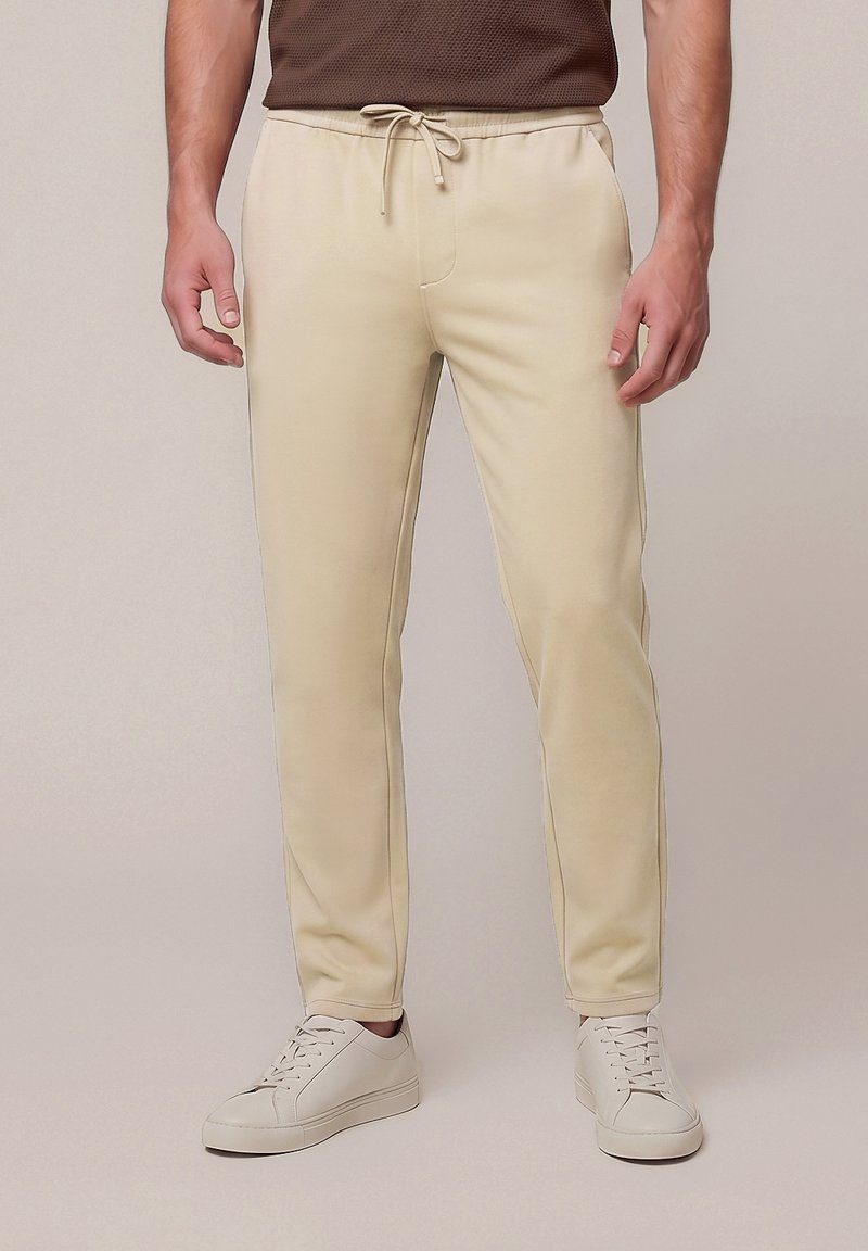 Homme portant un pantalon beige à cordon et des baskets blanches, debout devant un fond clair uni.
