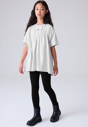 Gaiši pelēka pārmērīga izmēra T-krekls ar savilktu sānu detaļu, īsām piedurknēm un logotipu. Kombinēts ar melnām legingiem un melniem potītes garuma zābakiem.