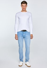 Jauns vīrietis stāv, tērpies baltā ar garām piedurknēm krekliņā, gaiši zilās džinsās, baltās sporta kurpēs un melnā jostasvietā pret vienkāršu gaišu fonu.