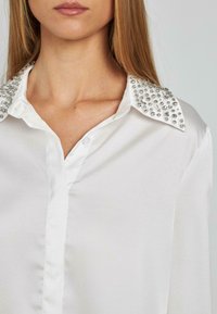 Camicia bianca con finitura in raso, dotata di un colletto adornato con strass trasparenti e chiusura frontale con bottoni.