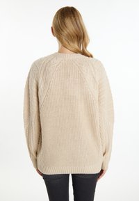 Pulover bej supradimensionat, tricotat, cu modele texturate de cablu, manșete cu căptușeală și un decolteu larg. Vedere din spate care arată croiala lejeră și detaliile de design.