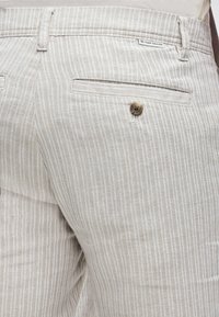 Pantalones a rayas finas beige y blancos con un bolsillo trasero abotonado y una pequeña etiqueta "Tom Tailor" encima del bolsillo.