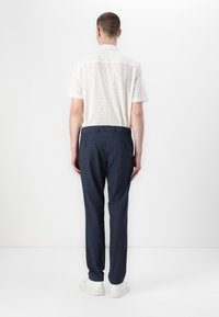 Hvit bluse med kort erme, marineblå bukser og hvite sneakers. Blusen har et teksturert mønster og standard krage, løs passform.