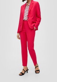 Completo su misura fucsia composto da una giacca con un solo bottone, pantaloni aderenti e una blusa a fantasia con accenti in bianco e nero.