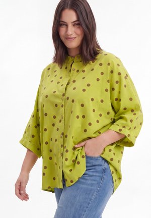 Smiling woman with shoulder-length dark hair wearing a loose lime green button-up shirt with brown polka dots and blue jeans.