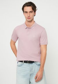 Νέος άνδρας με κοντά κυματιστά μαλλιά, ντυμένος με ανοιχτό ροζ πόλο μπλουζάκι και ανοιχτό μπλε τζιν, στέκεται μπροστά σε ένα απλό λευκό φόντο.