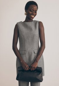 Pilkas be rankovių sijonas su tekstūruotu audiniu ir šoninėmis kišenėmis, papildytas juodu odiniu delnine, laikomu priešais.