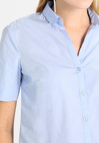 Chemise à manches courtes bleu clair à col pointu, en coton, avec une texture lisse et un léger éclat.