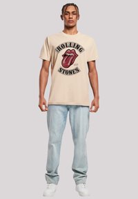 Beige katoen t-shirt met een rood en zwart Rolling Stones logo, gecombineerd met lichtgewassen jeans en witte sneakers. Casual pasvorm en ontwerp.