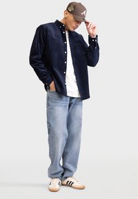 Marineblaues Cordhemd über einem weißen T-Shirt, kombiniert mit hellblauen, lockeren Jeans und cremefarbenen Sneakern mit schwarzen Streifen. Braune Mütze mit Logo.