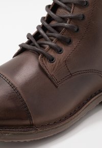 Hnědá kožená bota s hladkým povrchem, zaoblenou špičkou, tmavě šedými tkaničkami a viditelným šitím podél stran a podrážky.