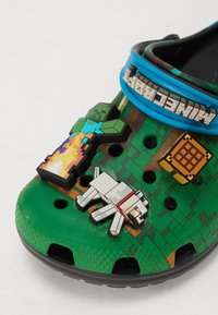 Gröna Crocs med pixelerade Minecraft-karaktärscharms: en zombie, en hund och ett hantverksbord. Har andningsbara hål och en blå kant.