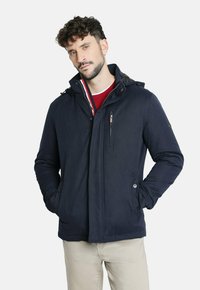 Chaqueta acolchada azul marino con un bolsillo en el pecho con cremallera, cierres de botón y capucha ajustable. Presenta un diseño elegante con una textura suave.