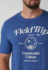 Blauwe katoenen t-shirt met ronde hals, met witte grafische tekst en een cirkelontwerp met afbeeldingen van vogels en landschappen.