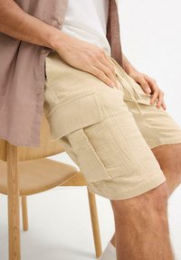 Beige Baumwoll-Cargo-Shorts mit Seitentaschen, mit einer strukturierten Oberfläche und einer lässigen Passform, kombiniert mit einem hellen Hemd, sitzend auf einem Holzstuhl.