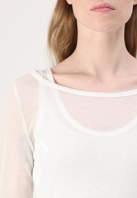 Hvit halvgenomsiktig langermet topp med rund halsutskjæring og subtil tekstur, med et lagdelt design nær kragen.