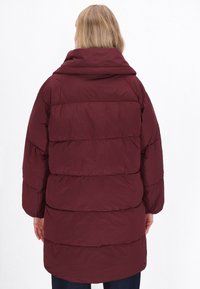 Giacca a vento bordeaux con cappuccio, caratterizzata da un design trapuntato e cuciture orizzontali. Tessuto morbido e maniche lunghe, che arrivano sino a metà coscia.
