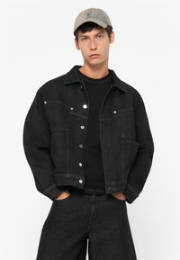 Joven con una gorra gris, chaqueta de mezclilla negra con botones plateados, camiseta negra y jeans negros, manos en los bolsillos, expresión neutral.