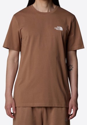 Osoba ubrana w brązowy T-shirt z krótkim rękawem i dopasowane spodnie z białym logo "The North Face" na lewej piersi oraz czarny tatuaż słońca na lewym przedramieniu.