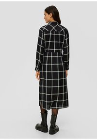 Schwarzes langärmliges Kleid mit weißem Karomuster, Knopfkragen und gerafftem Rückendetail; kombiniert mit schwarzen klobigen Knöchelstiefeln.