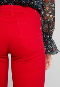 Los jeans rojos presentan un diseño ajustado y suave con bolsillos traseros y sutiles acentos de costura. La tela tiene un color sólido y vibrante con un ligero brillo.